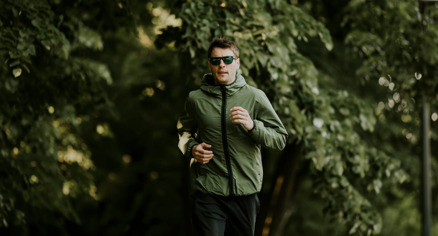 Man jogging outdoors in a green jacket and dark sunglasses, surrounded by lush green trees, representing an active and eco-conscious lifestyle.